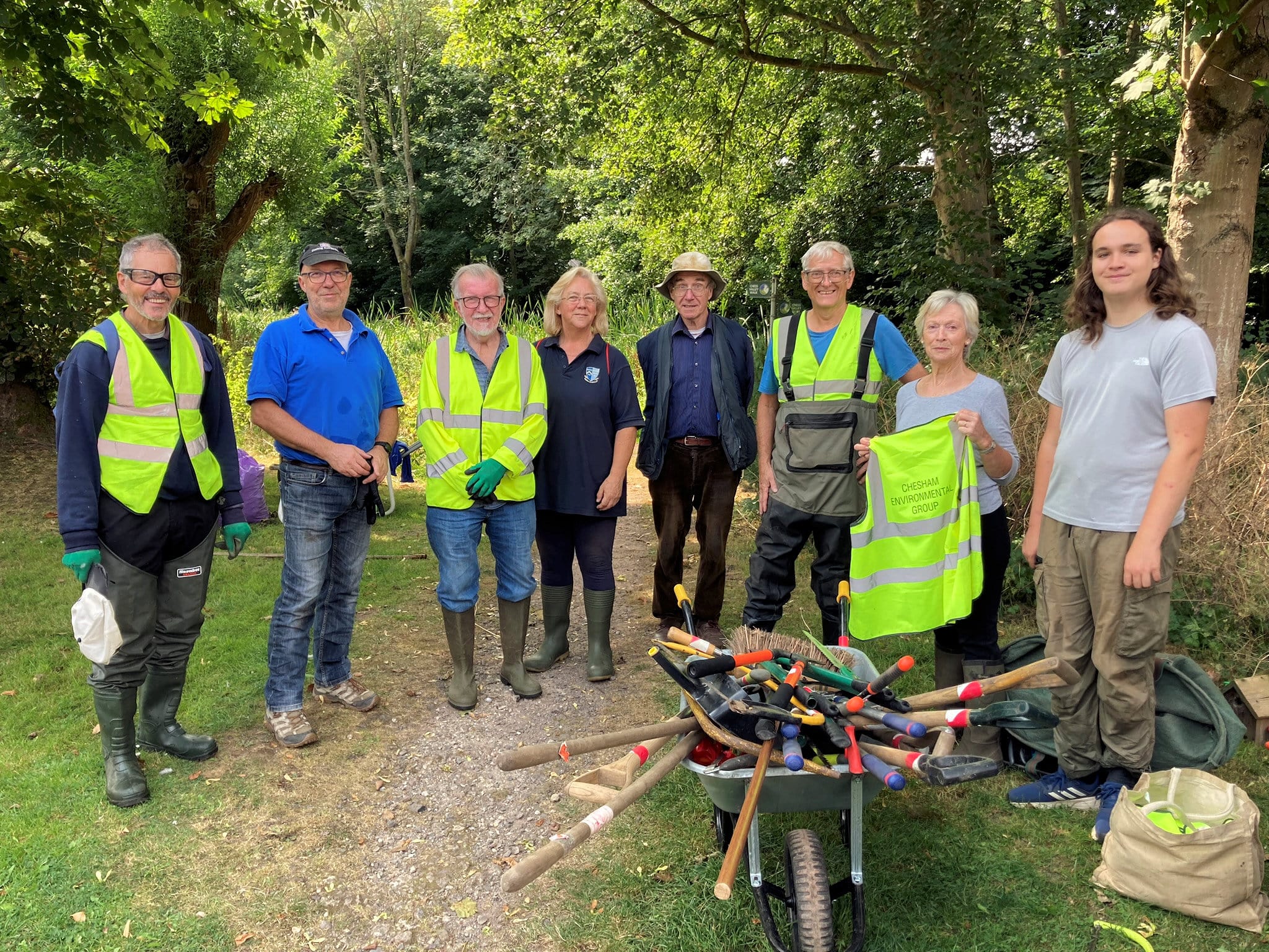 A group of eight volunteers posing outdoors in a green, wooded area during daytime. Some are wearing high-visibility vests, gloves, and boots, while others are in casual clothing. They are standing on a dirt path with a wheelbarrow in front of them, filled with various gardening tools such as rakes, trowels, and spades. One volunteer is holding a high-visibility vest with the words 'Chesham Environmental Group' printed on it. The group are engaged in an environmental or community cleanup activity.